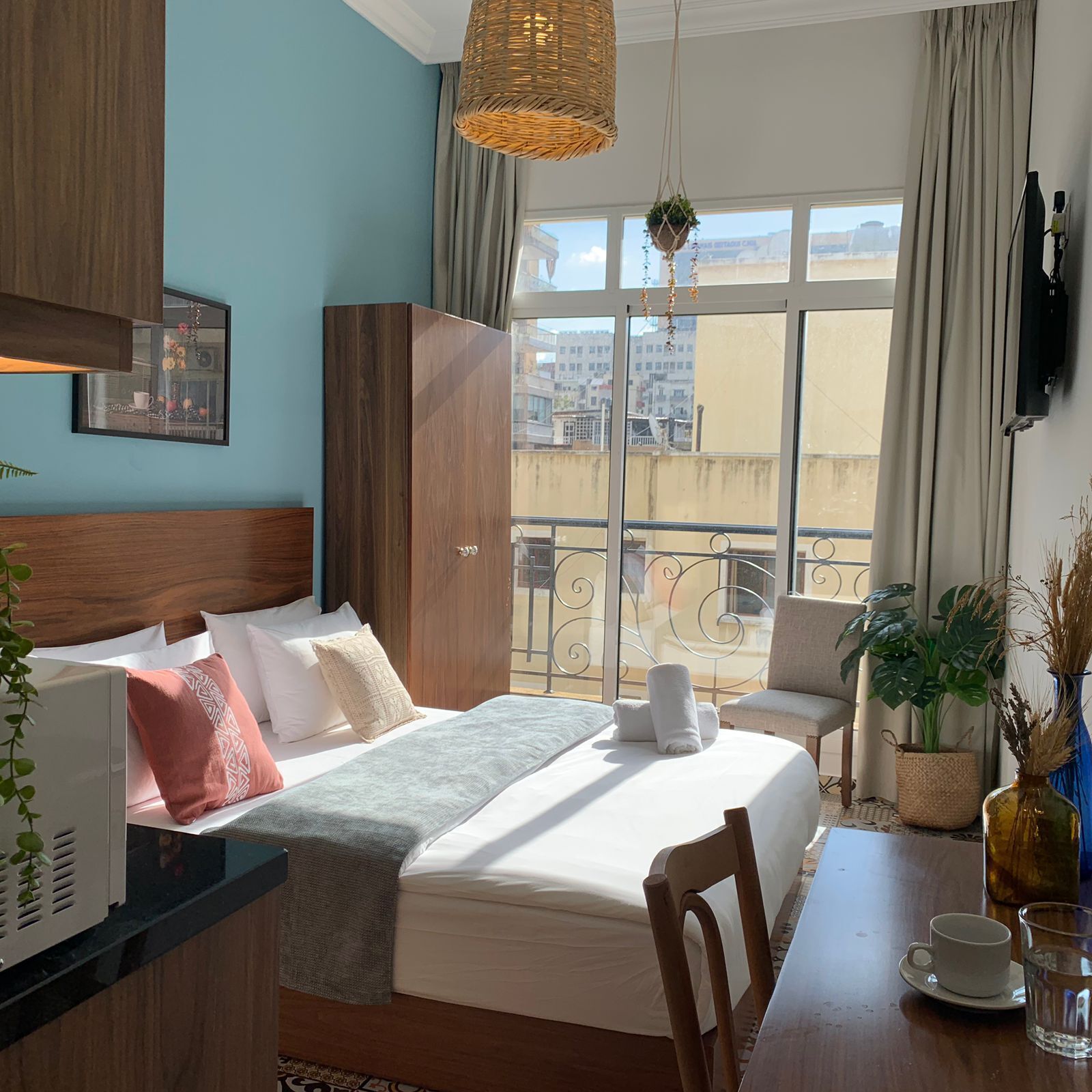Ett hotellrum med en säng, ett skrivbord, en stol, ett fönster med utsikt över byggnader och en hängande växt.