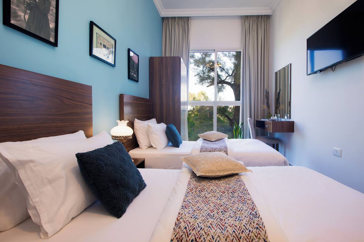 A hotel room with two beds, a TV, and a window with a view of trees.