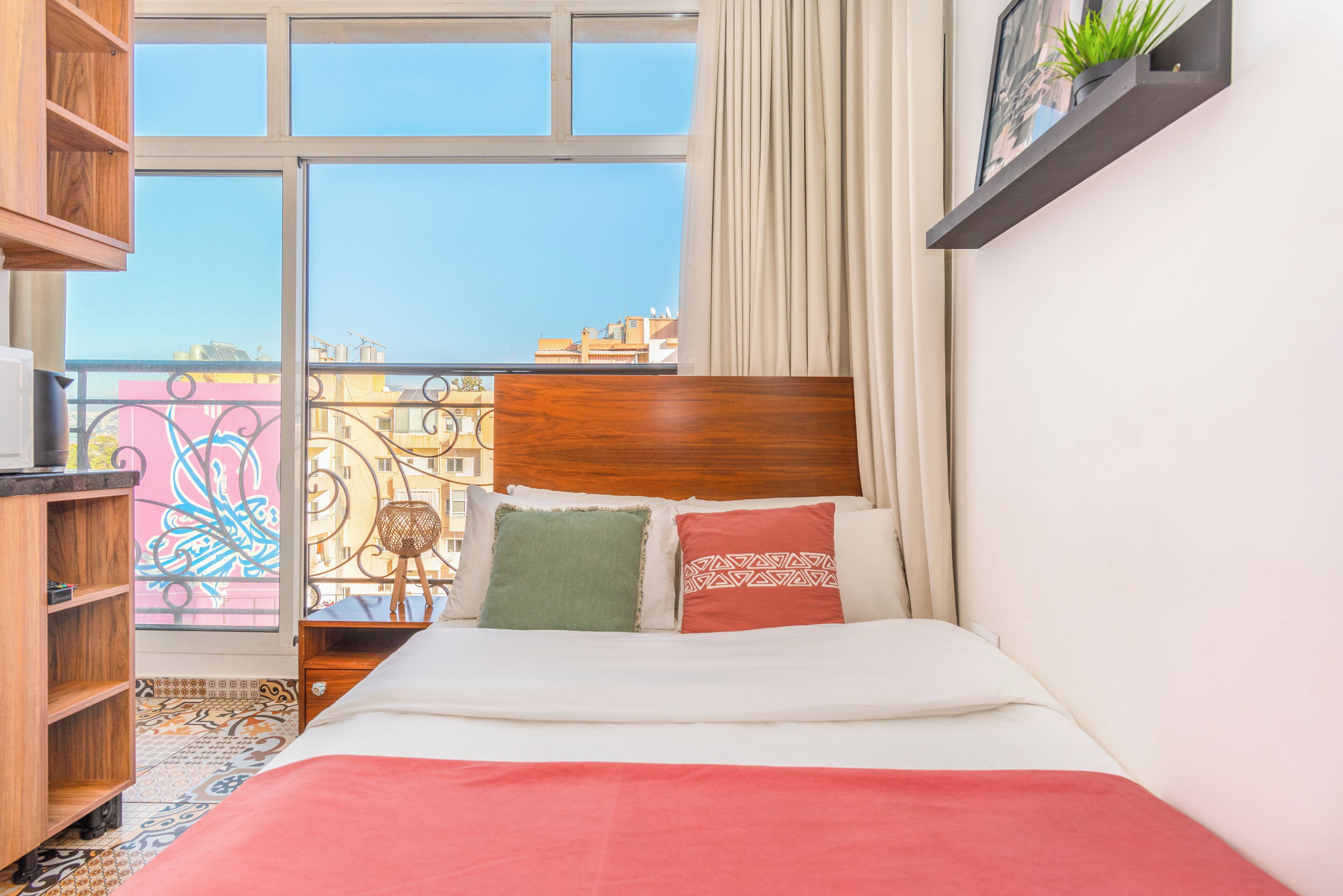 A hotel room with a bed, a wooden nightstand, a shelf with a plant, and a view of buildings through a window.