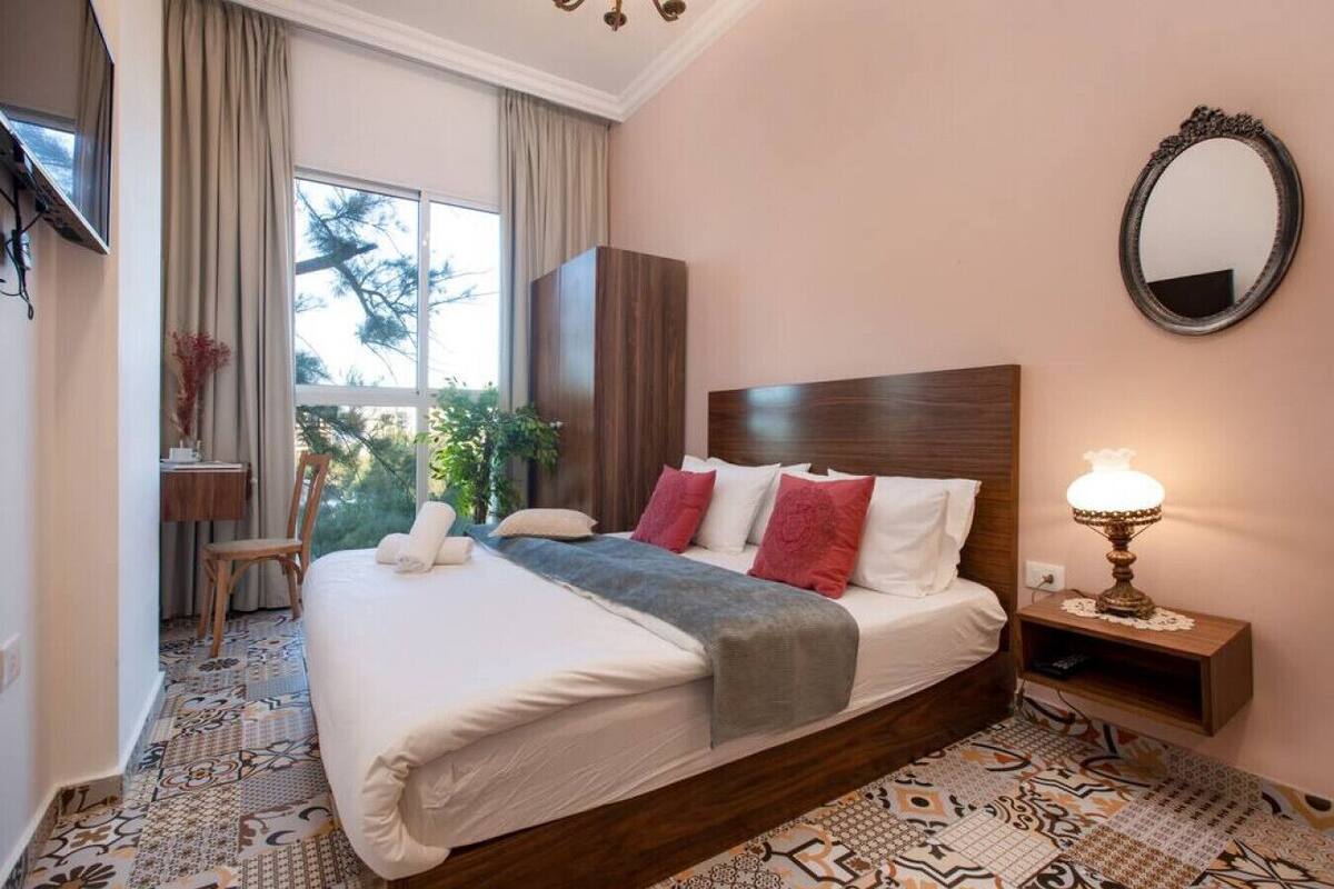 A hotel room with a large bed, a desk, a chair, a bedside table with a lamp, and a window with curtains.
