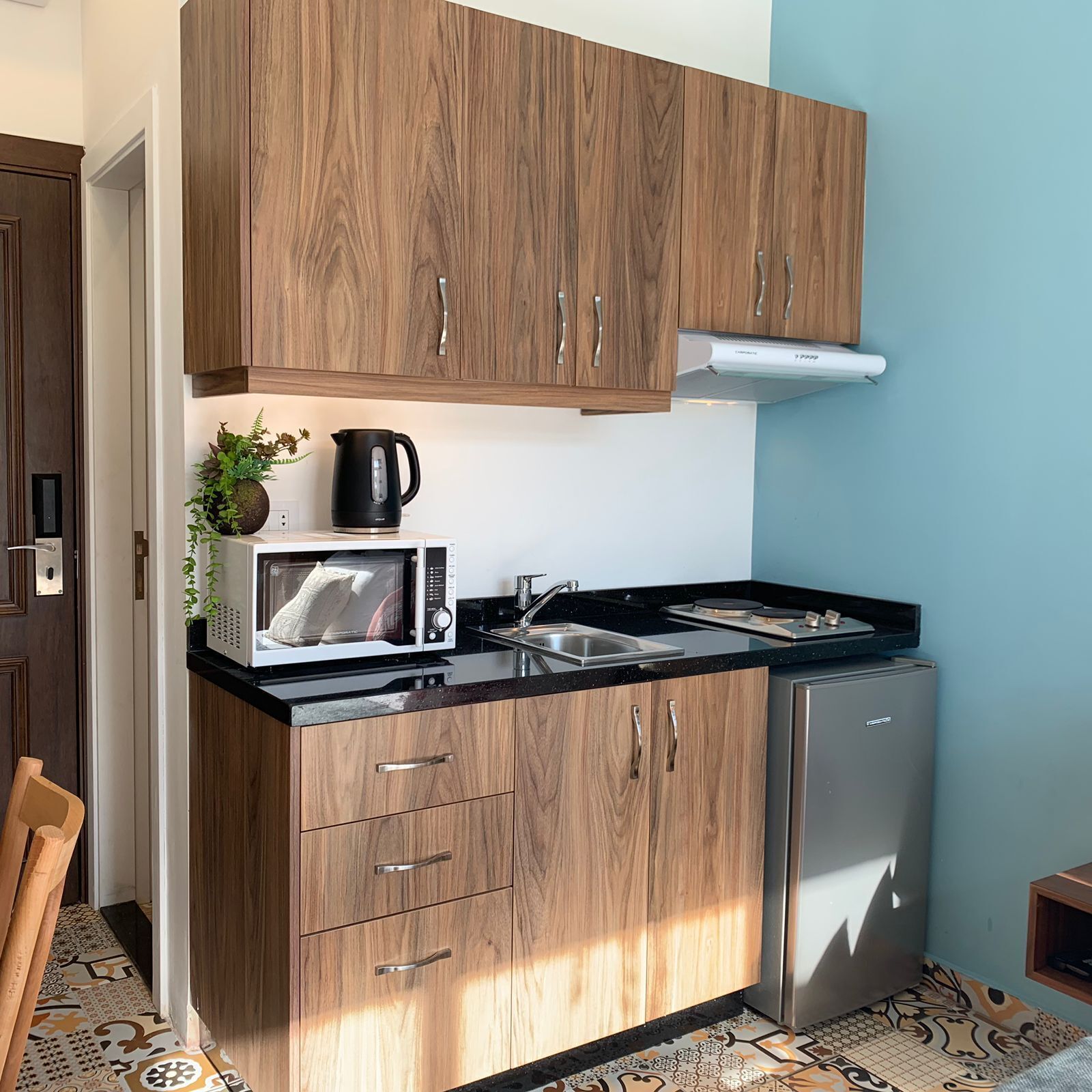 A compact kitchen with wooden cabinets, a microwave, a kettle, and a stainless steel refrigerator.