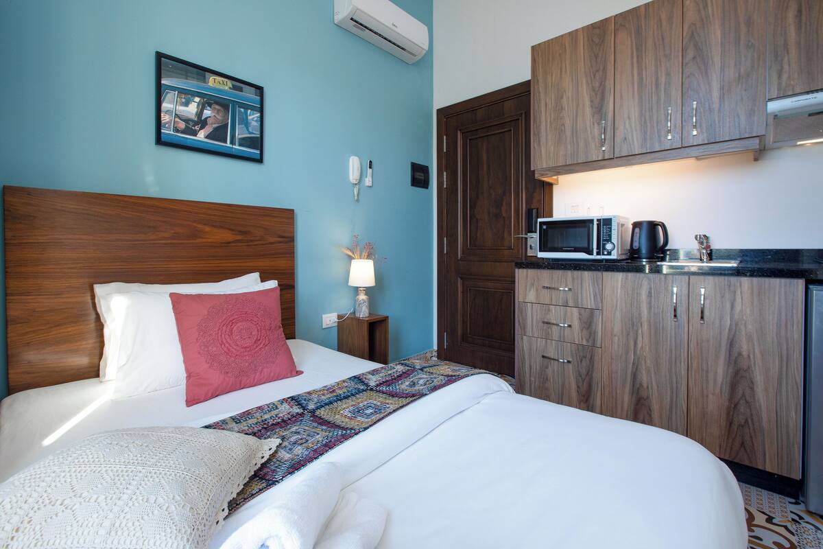 A hotel room with a bed, a wooden wardrobe, a microwave, and a small table with a lamp.