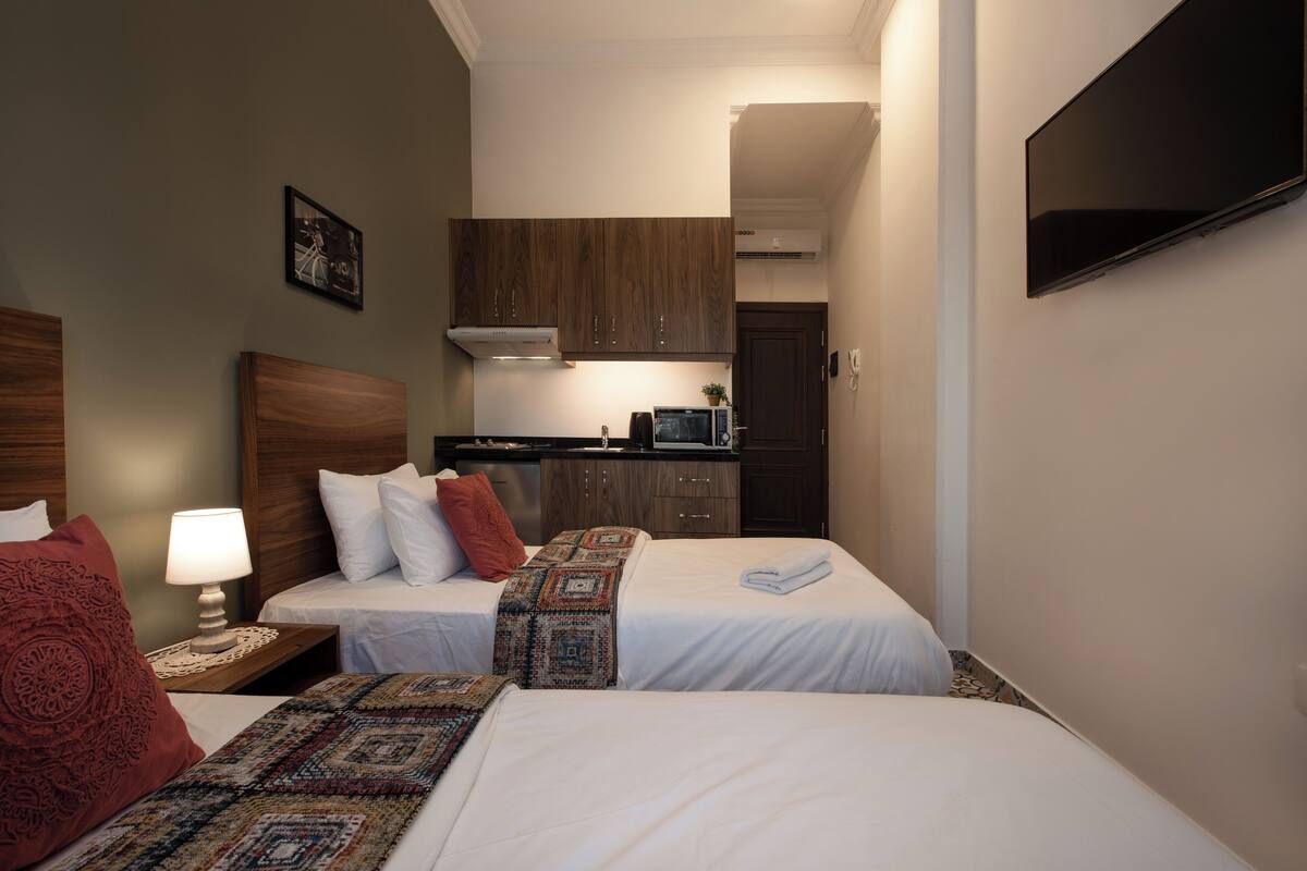 A hotel room with two beds, a small kitchenette, and a TV mounted on the wall.