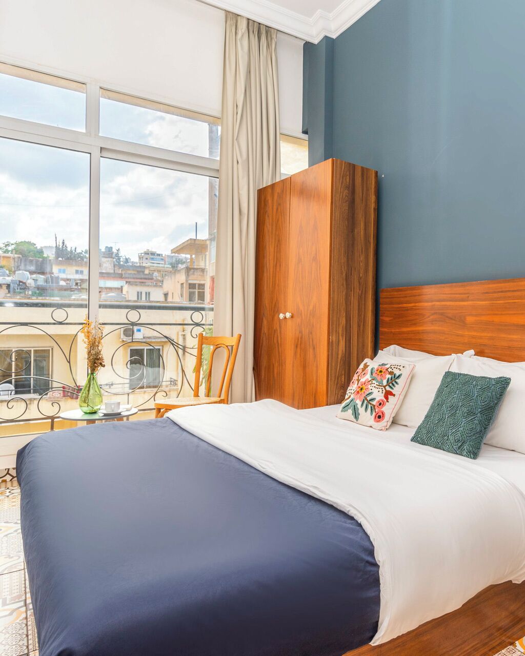 Habitación de hotel con una cama grande, un armario de madera, vista de la ciudad a través de un ventanal y un balcón con una silla y un jarrón.