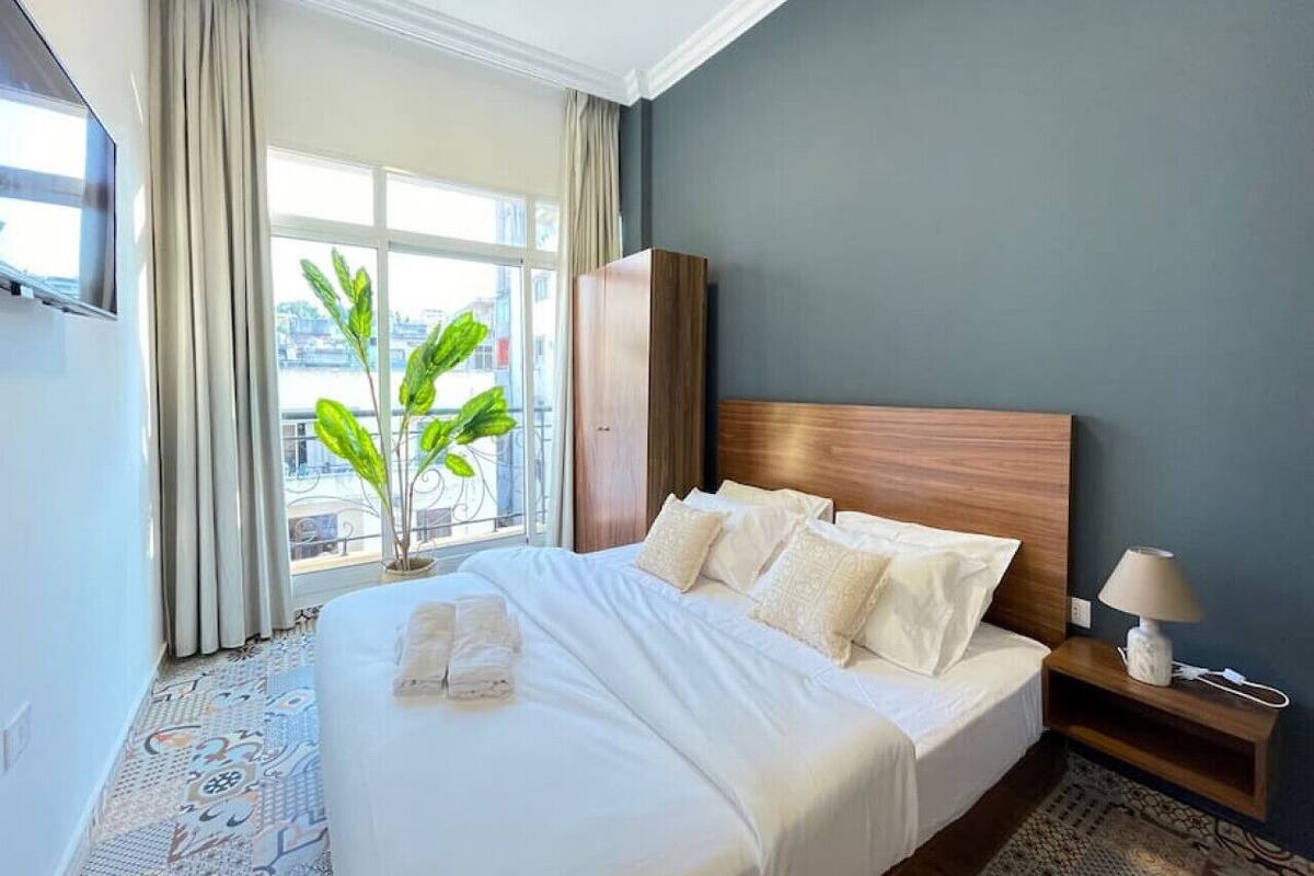 A hotel room with a large bed, a nightstand with a lamp, a wardrobe, and a view of the city through the window.