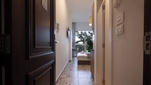 A hallway with patterned floor tiles, a bed with a view of trees, and a door with a light switch. - Mar Mikhael Studios (Beirut)