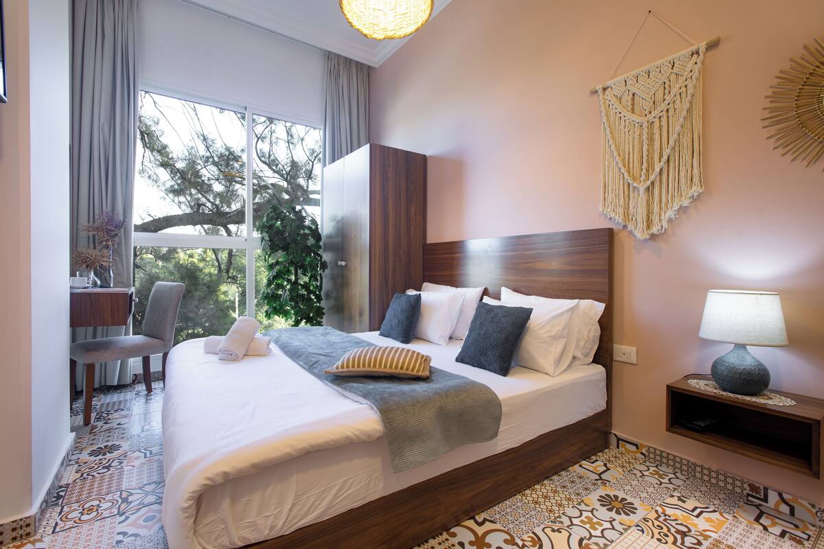 A hotel room with a large bed, a desk with a chair, a window with curtains, and a wall-mounted lamp.