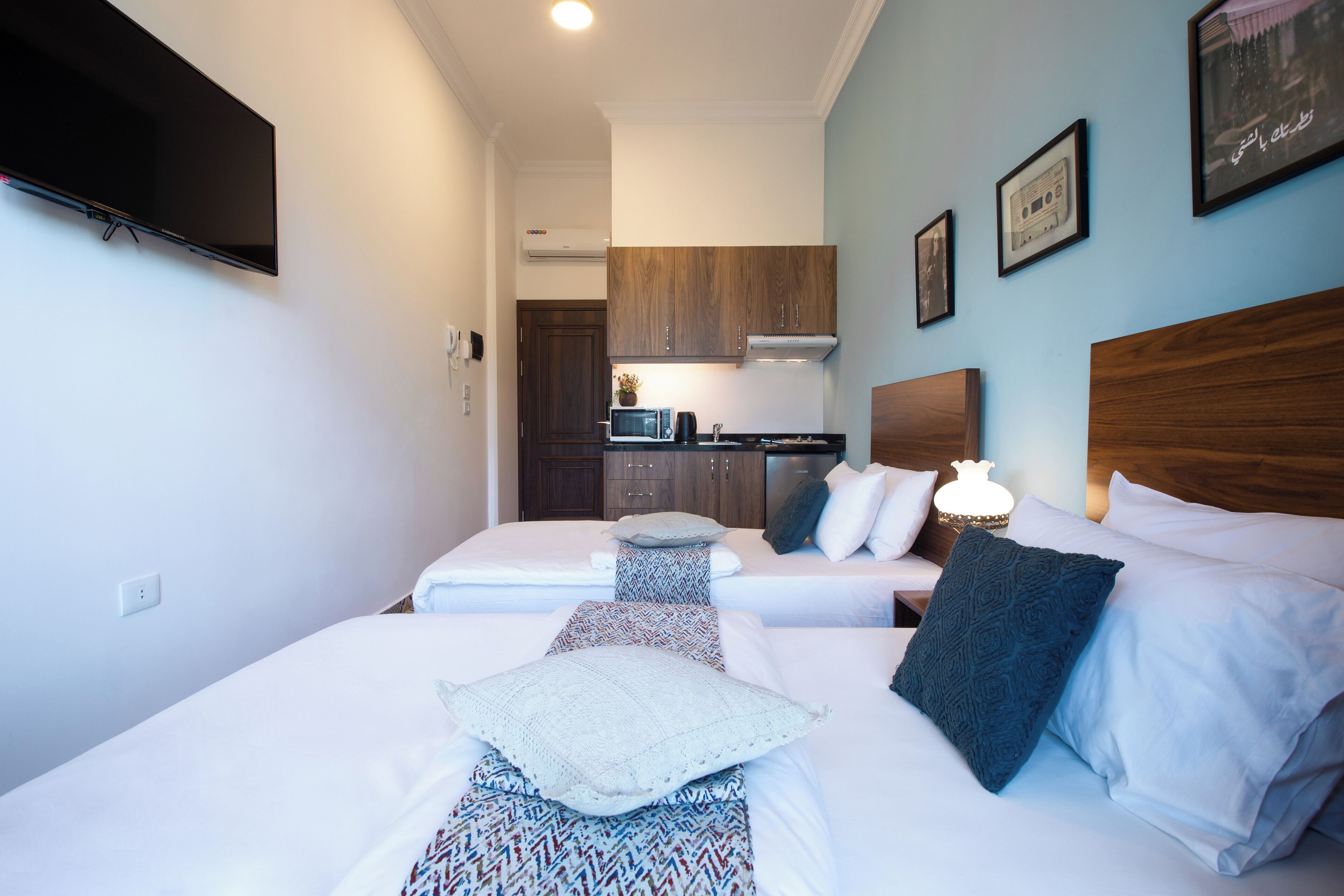 Habitación de hotel con dos camas, kitchenette y televisor montado en la pared.