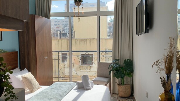A hotel room with a bed, a desk with a cup, a TV, a balcony with a view of buildings, and a hanging plant. - Mar Mikhael Studios (Beirut)