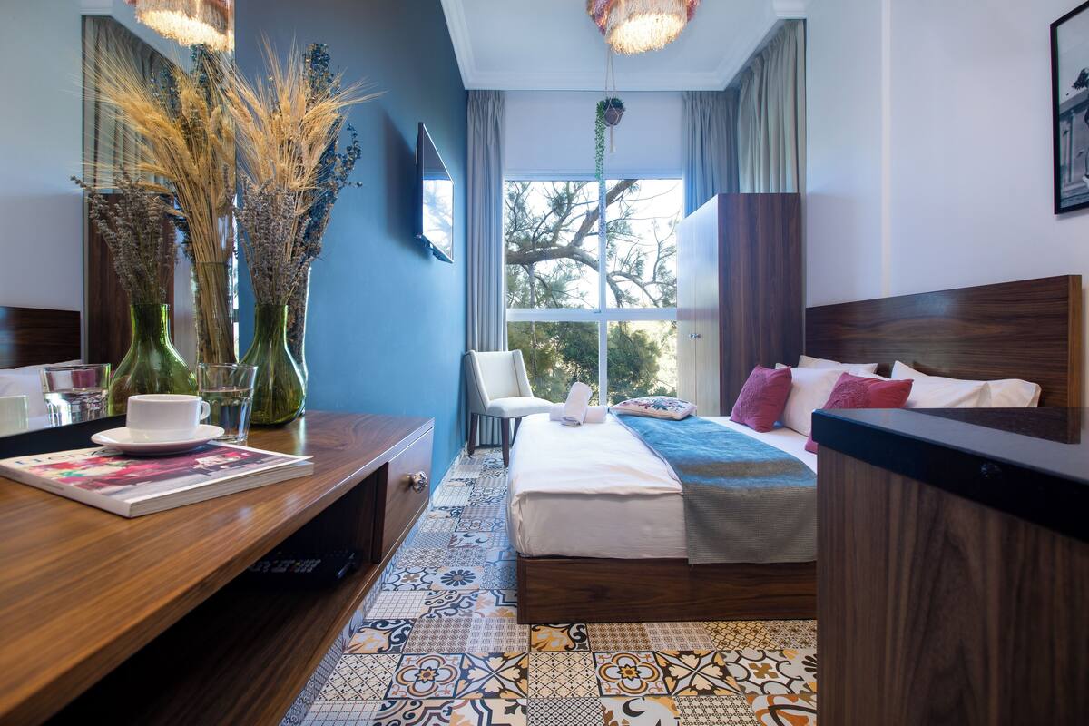 A hotel room with a bed, bedside table, and a window with a view of trees.