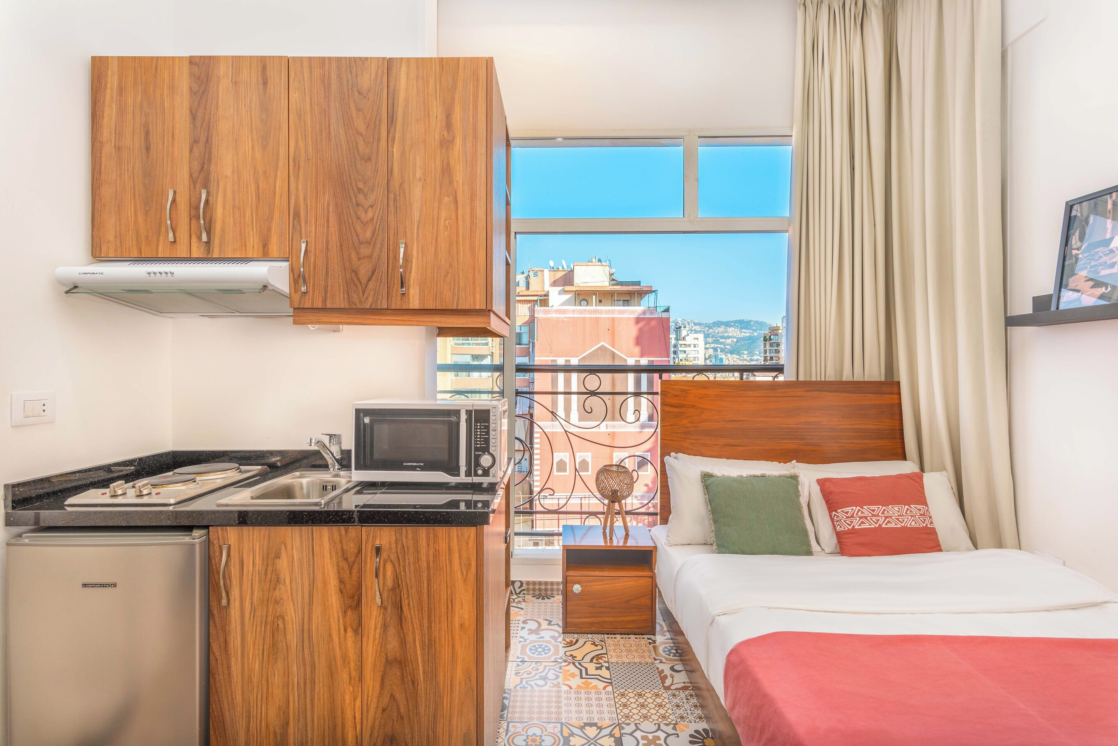 A compact hotel room with a kitchenette, a bed with a wooden headboard, a view of the city through large windows, and a patterned floor.