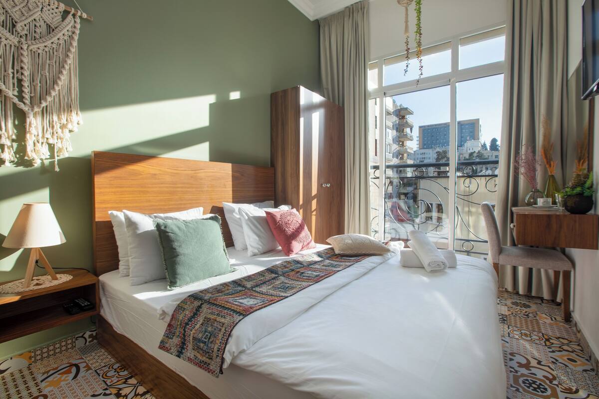 A hotel room with a large bed, a wooden wardrobe, a nightstand with a lamp, and a view of the city through the window.