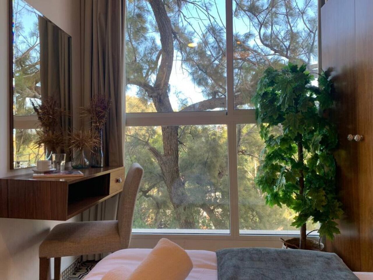A bedroom with a large window, a desk with a mirror, a chair, a bed, and a potted plant.