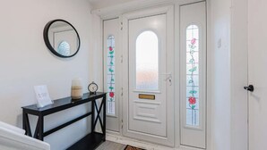 Un pasillo con una puerta blanca que tiene un panel de vidrio esmerilado con diseños florales, una consola negra con un espejo, un reloj y un jarrón pequeño.