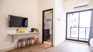 A modern room with a flat-screen TV, a small desk with a chair, a bathroom visible through a glass door, and a large window with a view of buildings. - BAOBAOB&B (Magong)