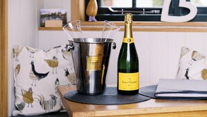 A cozy room with a wooden table, a champagne bottle, and a bird-themed cushion. - Shepherd's Hut- Hot Tub- Wellness Spa- Wood Burner (Nottingham)