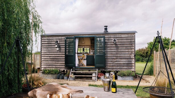 A rustic wooden cabin with a green door, a table with a bottle, and a metal bucket. - Shepherd's Hut- Hot Tub- Wellness Spa- Wood Burner (Nottingham)