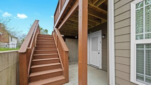 Eine Holztreppe führt zu einem überdachten Bereich mit einer weißen Tür und einem Fenster mit Jalousien.