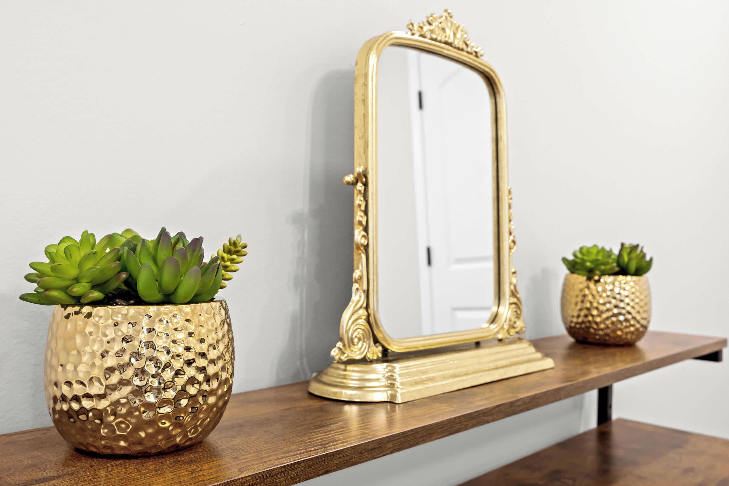 A gold-framed mirror on a wooden shelf with two gold textured pots containing succulents.