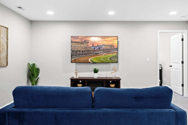Un salón con un sofá azul, un televisor de pantalla plana montado en la pared, una planta pequeña en el mueble de televisión y una puerta a la derecha.
