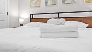 Ein ordentlich bezogenes Bett mit weißen Leinen und aufgerollten Handtüchern, ein Holz-Kopfbrett und drei gerahmte Kunstwerke an der Wand.