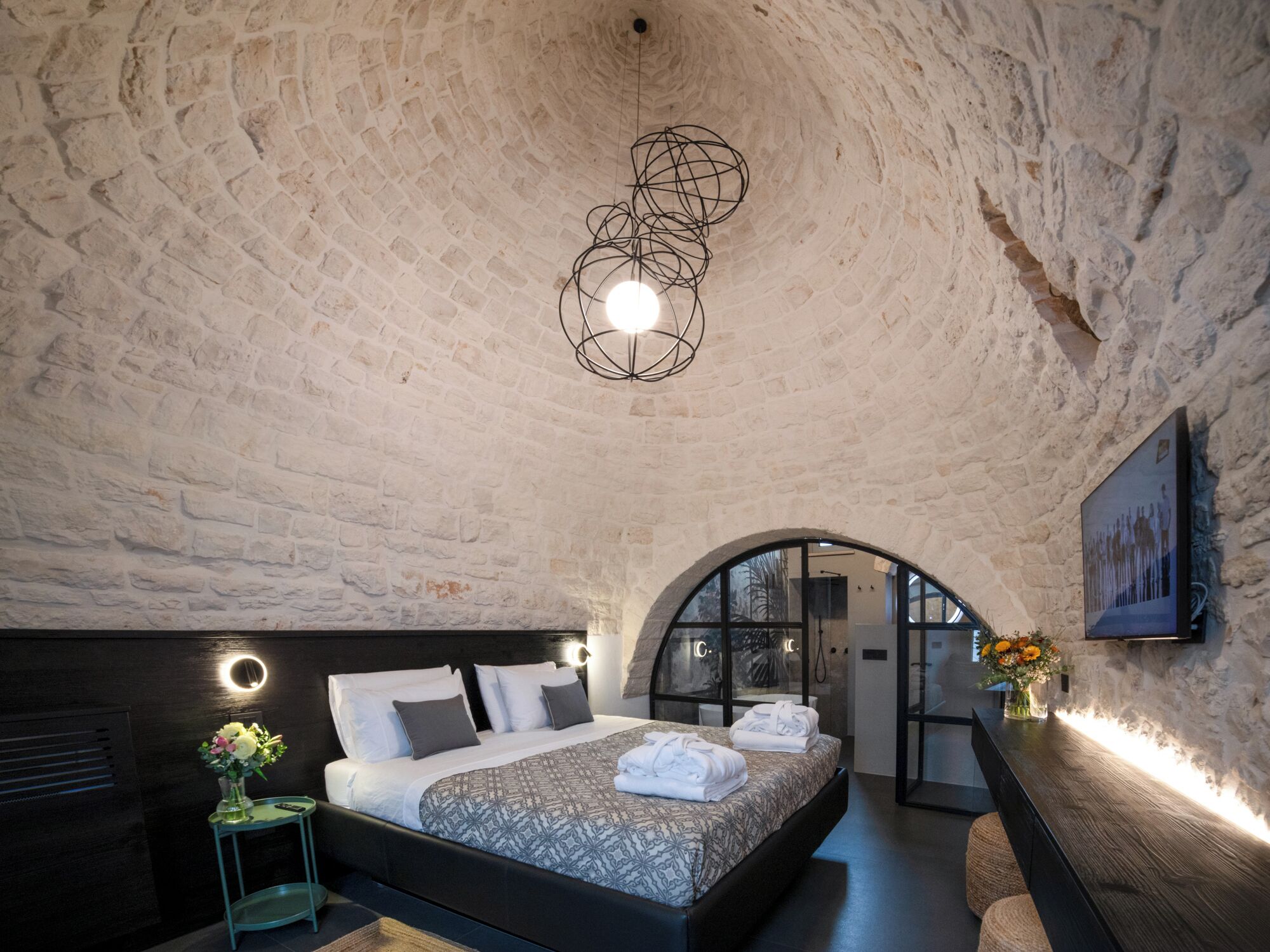 A bedroom with a stone wall, a modern bed, a glass door leading to a bathroom, and a pendant light.