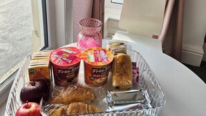 A tray of food items including pastries, a jar of jam, and a bottle of syrup on a table by a window with curtains.