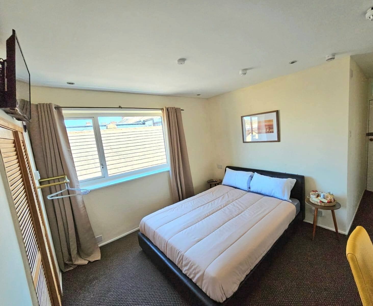 A hotel room with a bed, a small table with a chair, a window with curtains, and a picture on the wall.