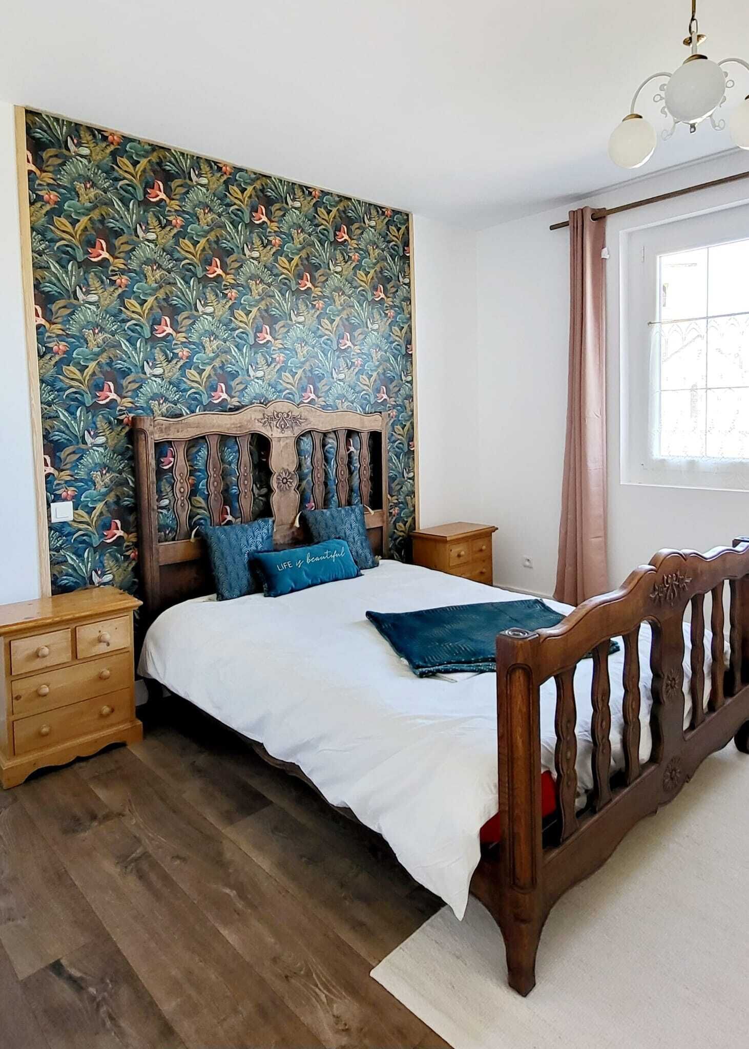 Ein Schlafzimmer mit einem hölzernen Bett, einem Kopfstück mit Blumenmuster, einem Nachttisch und einem Fenster mit Vorhängen.