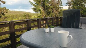 Une table ronde avec une tasse blanche et deux plantes en pot, une clôture en bois sombre et une vue sur un paysage boisé.