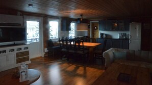 Een ruime woonkamer met houten plafond, een eettafel met stoelen, een bank en een keuken met kasten en apparatuur.