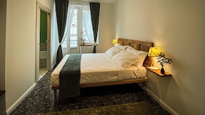 A neatly made bed with a wooden headboard, a bedside table with a lamp, and a view of a balcony through sheer curtains. - Caramia srls (Crispiano)