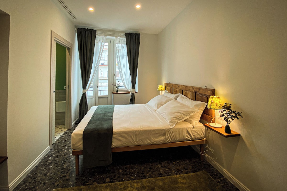 Una cama bien hecha con cabecera de madera, una mesita de noche con lámpara, y vista a un balcón a través de cortinas traslúcidas.