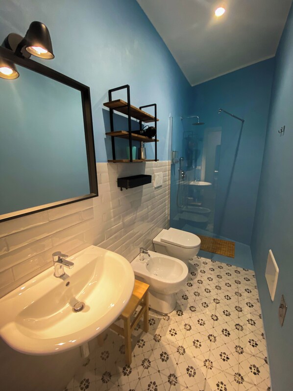 A bathroom with a white sink, a bidet, and a toilet. There is a mirror above the sink, and a shelf with a black frame on the wall. The floor has a patterned tile design. - Caramia srls (Crispiano)