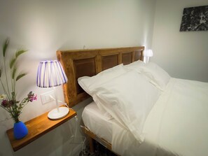 A neatly made bed with white linens, a wooden headboard, a bedside table with a lamp, and a vase with flowers. - Caramia srls (Crispiano)