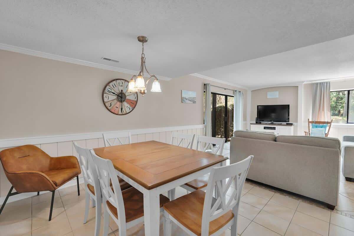 A dining area with a wooden table and white chairs, a sofa, a TV, and a clock on the wall.
