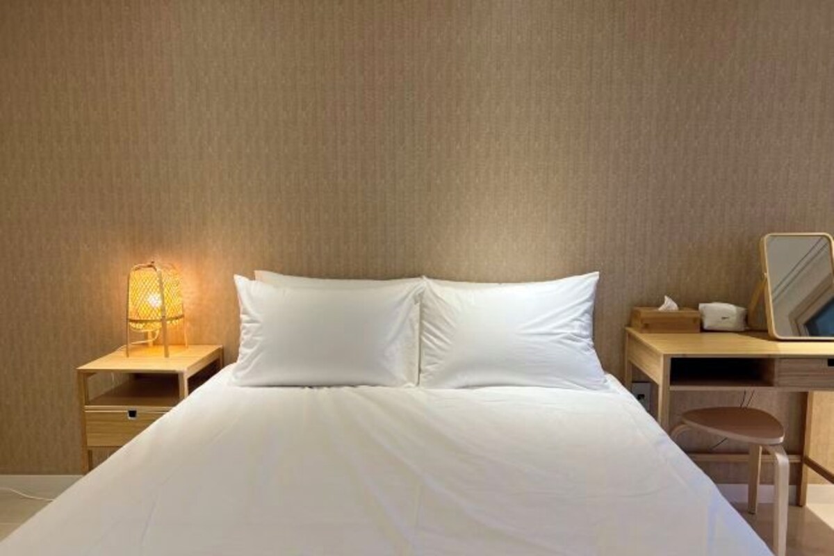 A neatly made bed with white linens, a bedside lamp, a wooden nightstand, and a desk with a chair.