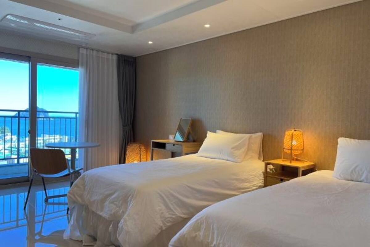 A hotel room with two beds, a small table, a chair, and a balcony with a view.