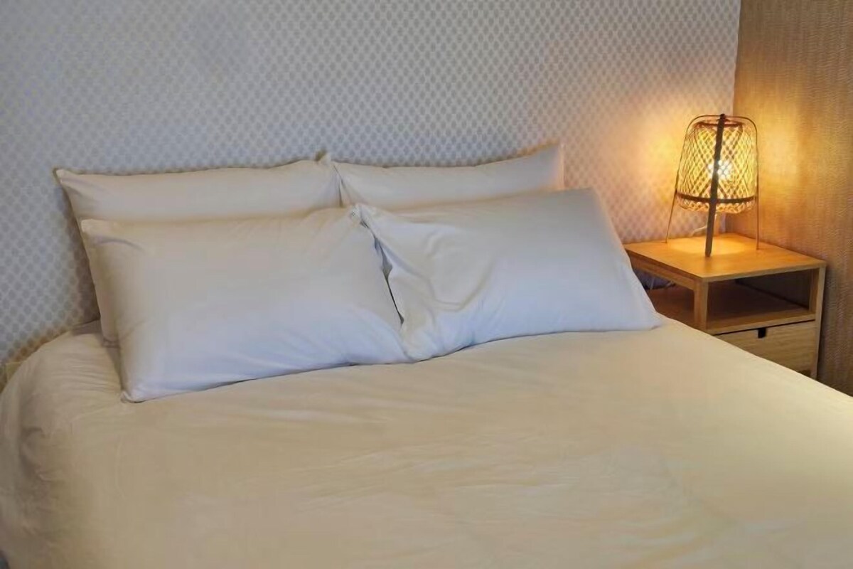A neatly made bed with white linens and pillows, a wooden nightstand with a lamp, and a patterned wall.