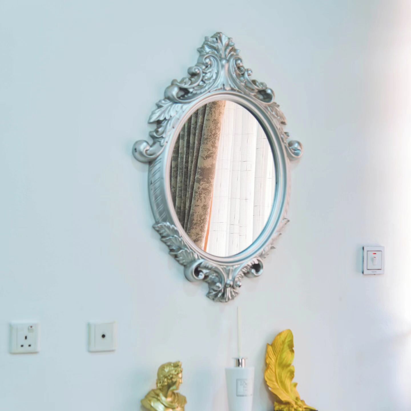 A mirror with an ornate frame, a gold statue, a white bottle, and a light switch on the wall.