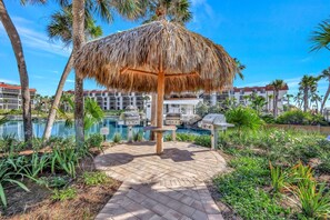 A pool area with a thatched umbrella, palm trees, and a barbecue grill.