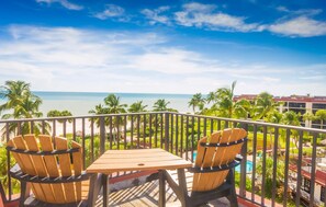 Ein Balkon mit zwei Holzstühlen und einem Tisch, mit Blick auf einen Strand und einen Poolbereich.