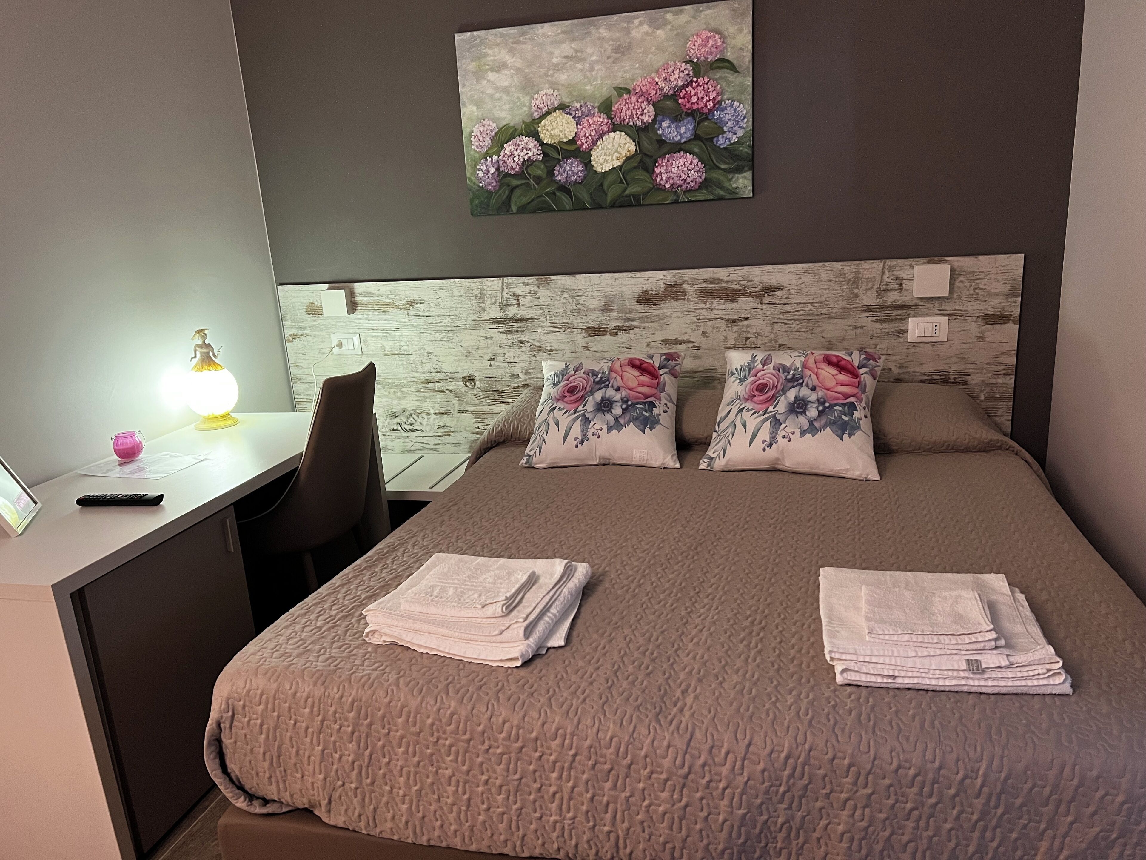 Ett välordnat hotellrum med en säng, ett skrivbord och en stol. På väggen ovanför sängen hänger en tavla med ett blommotiv.