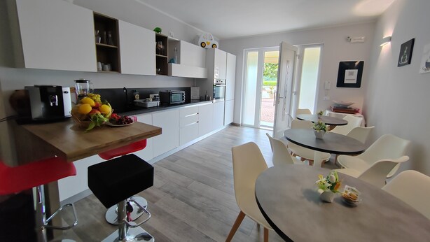A modern kitchen with white cabinets, a dining area with a round table and chairs, and a view through the open door to the outside. - Charming 4-bedroom estate in enchanting Albaré Stazione with AC, WiFi (Albaré Stazione)