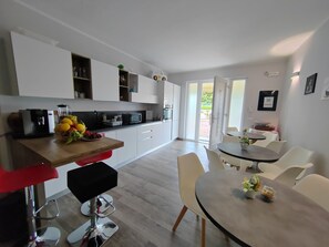 A modern kitchen with white cabinets, a dining area with a round table and chairs, and a view through the open door to the outside.
