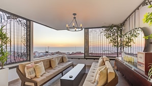 Una sala moderna con un gran ventanal que ofrece vistas al mar y al paisaje urbano al atardecer.