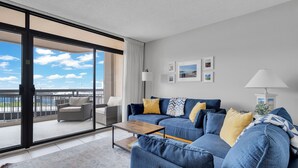 A modern living room with a blue sofa, yellow cushions, a wooden coffee table, and a view of the sky and balcony. - Perfect beach getaway at Fountainhead Towers, located oceanfront on 116th Street (Ocean City)