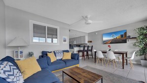 A modern living room with a blue sofa, a wooden coffee table, and a dining area with white chairs and a wooden table. - Perfect beach getaway at Fountainhead Towers, located oceanfront on 116th Street (Ocean City)