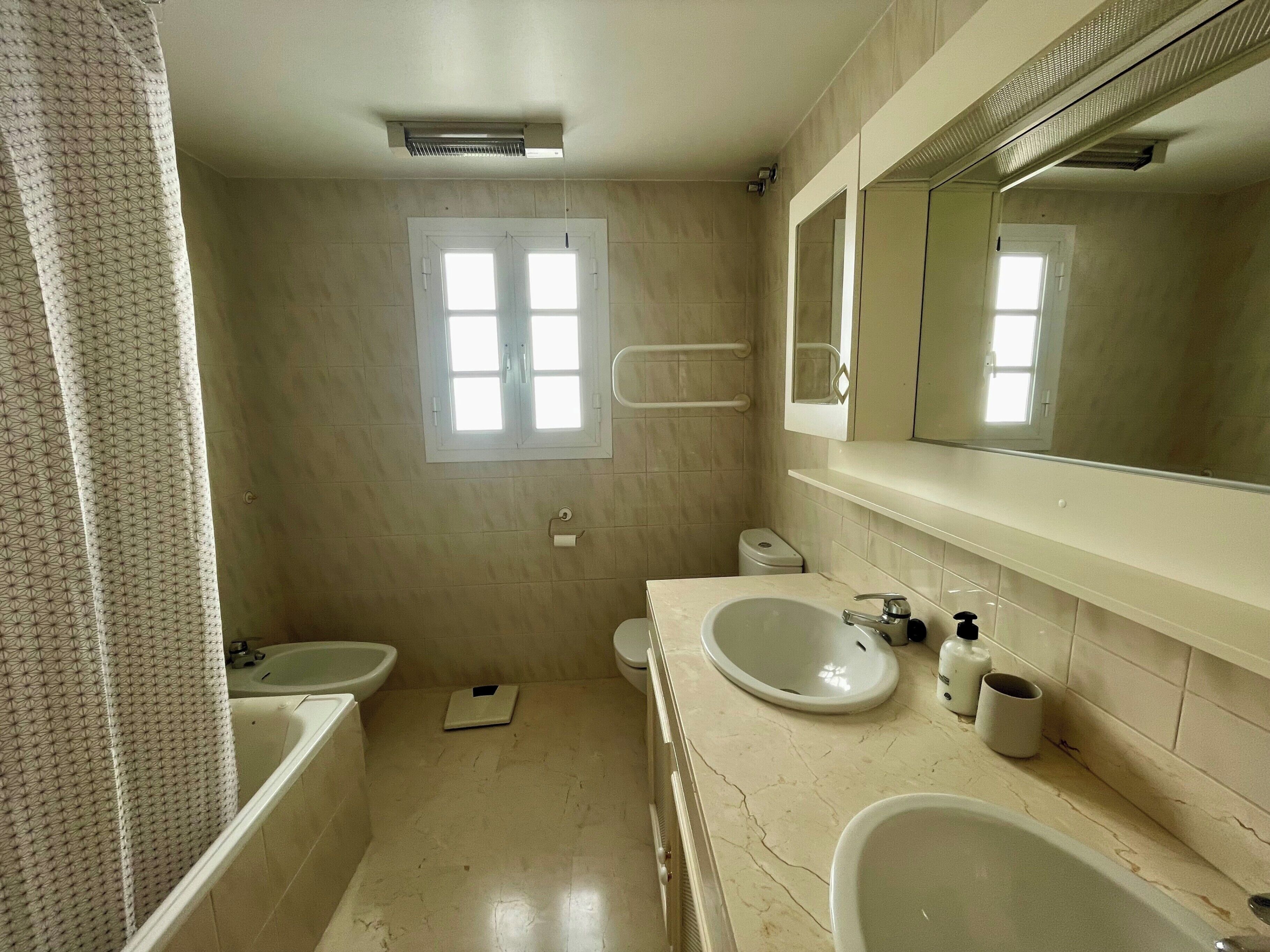 Ein Badezimmer mit Toilette, Waschbecken und Badewanne. Über der Toilette befindet sich ein Fenster, über dem Waschbecken ein Spiegel.
