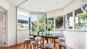 A modern kitchen with a glass dining table, wooden stools, and large windows offering a view of the garden. - Fairview Villa by Cadenzalux (Cascais)